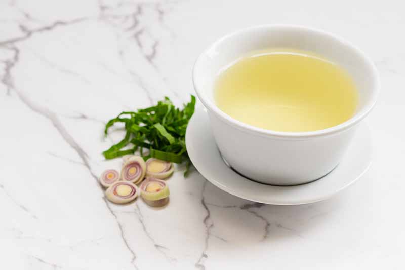 نمای نزدیک از یک فنجان و نعلبکی چینی سفید با یک چای زرد رنگ.  در سمت چپ فنجان تکه‌هایی از برگ‌ها و ساقه‌های علف لیمو، روی سطحی از سنگ مرمر قرار دارد.