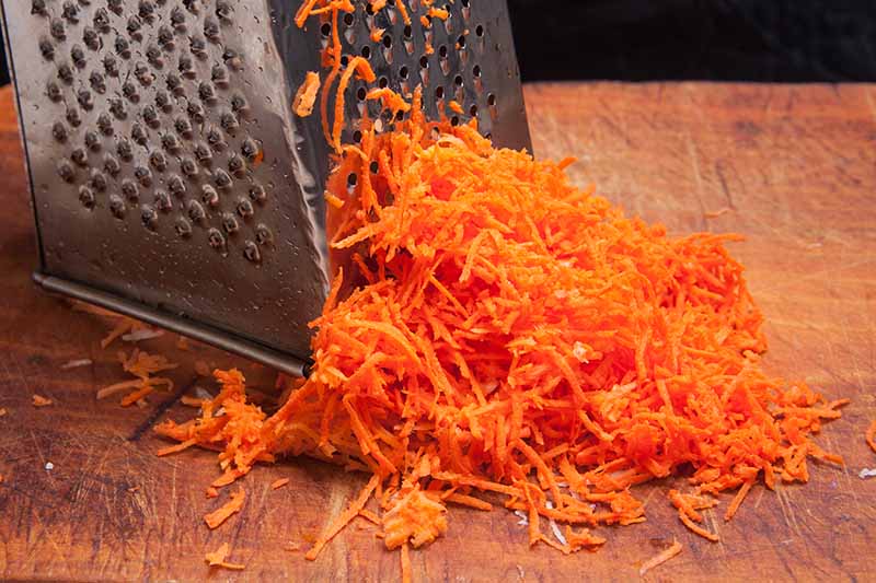 نمای نزدیک از یک رنده پنیر که برای رنده کردن هویج روی تخته خردکن چوبی روی زمینه تیره استفاده می شود.