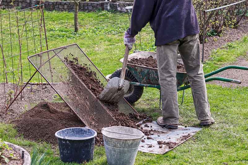 مردی که خاک باغچه را از چرخ دستی بیرون می آورد و آن را از طریق شبکه فلزی الک می کند تا کلوخه ها و سنگ ها را جدا کند.  در پس زمینه یک صحنه باغ است.