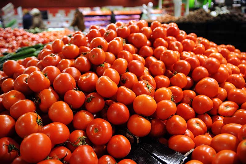 نمایشگر بزرگ سوپرمارکتی از گوجه‌فرنگی‌های قرمز رسیده، با خریداران و کابینت‌ها در فوکوس ملایم در پس‌زمینه.