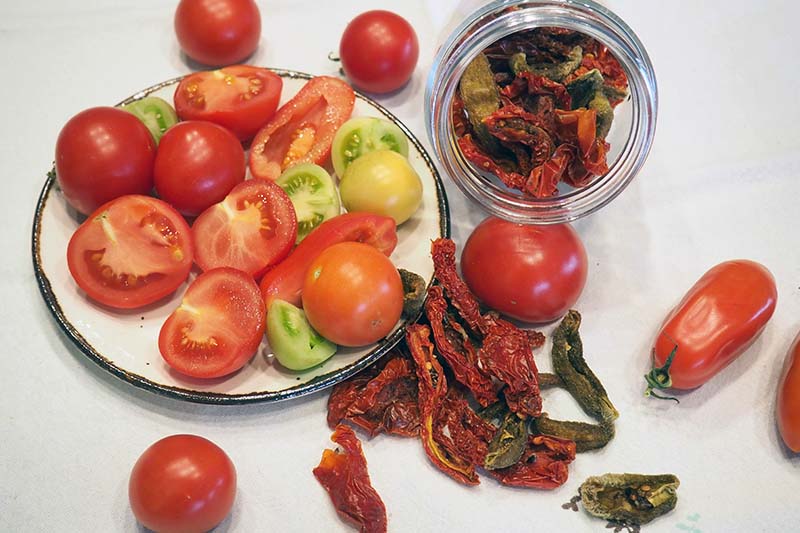 تصویر افقی نزدیک از یک بشقاب از گوجه‌فرنگی‌های تازه برداشت شده در سمت چپ قاب و گوجه‌فرنگی‌های آب‌گرفته در شیشه‌ای در سمت راست، روی یک سطح سفید قرار گرفته‌اند.