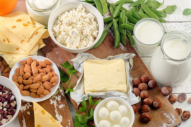 تصویر افقی نزدیک از انواع غذاهای مختلف: آجیل، پنیر، شیر و سبزیجات روی تخته خردکن چوبی.