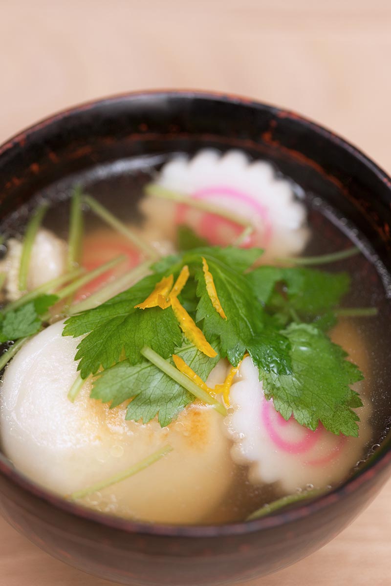 تصویر عمودی نزدیک از یک سوپ شفاف با کوفته‌های تزئین شده با میتسوبا که در پس‌زمینه‌ای با فوکوس ملایم تصویر شده است.